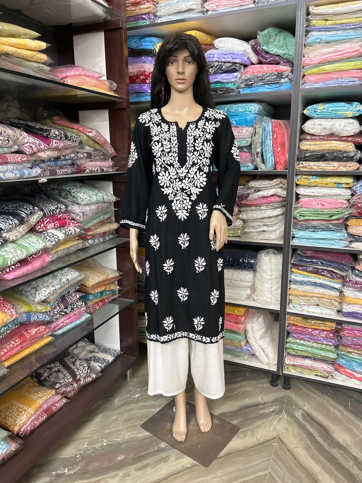 lucknowi chikankari Mannequin wearing a black and white patterned dress in front of shelves filled with colorful fabric.
