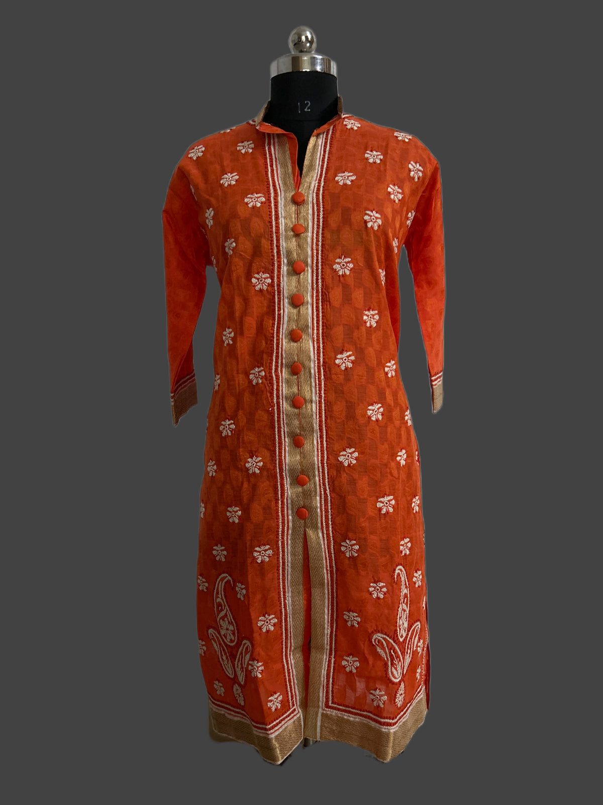 Lucknowi chikankari An orange long kurta with white floral prints and beige borders, displayed on a black dress form mannequin against a dark gray background.