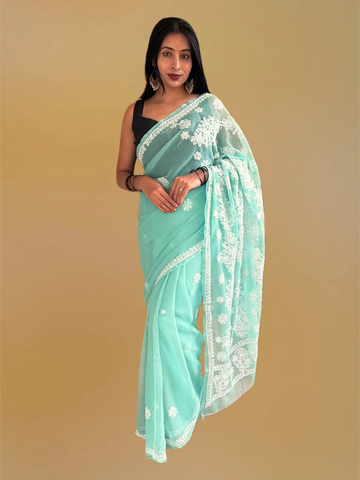 A woman modeling a mint green saree with white floral embroidery and a black sleeveless blouse, set against a solid beige background.