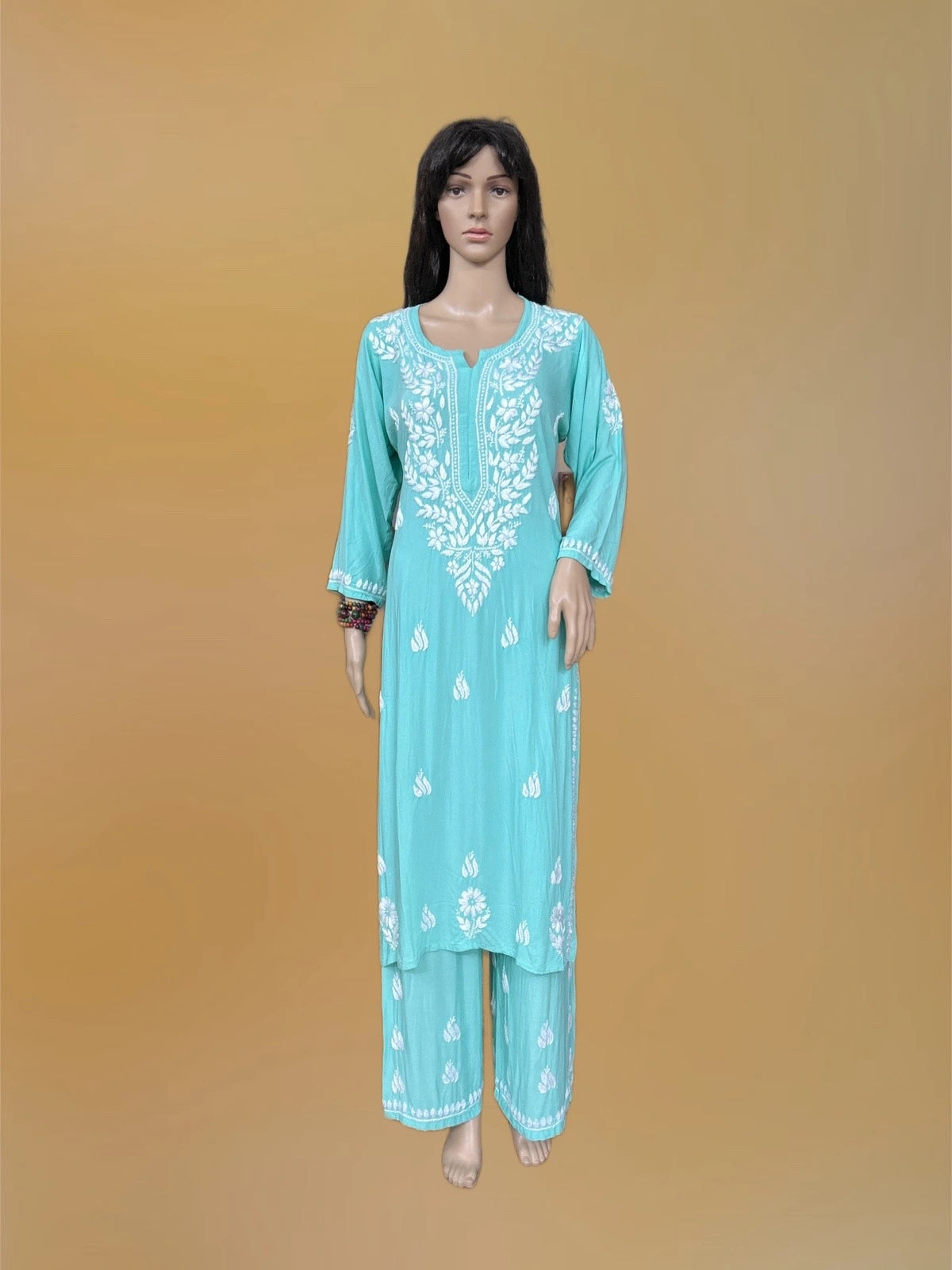A mannequin wearing a turquoise blue traditional tunic and trouser set with white floral embroidery, set against a solid tan background.