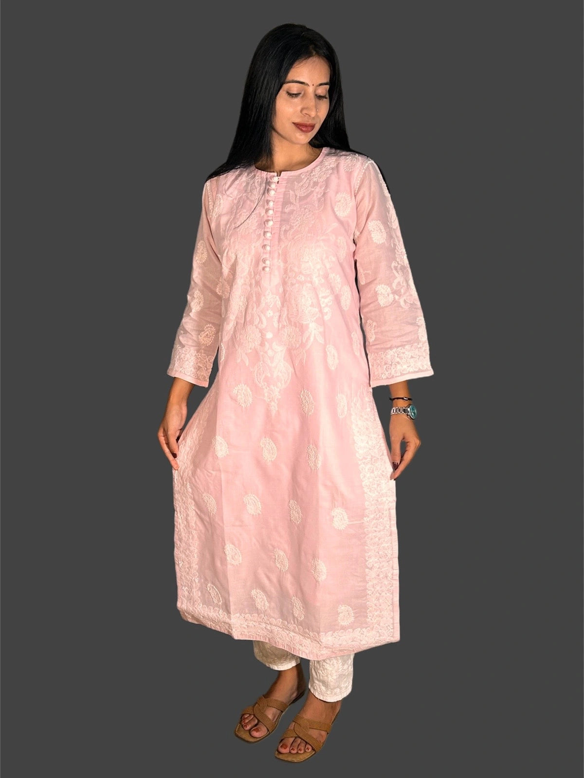 A woman modeling a light pink embroidered kurta with matching light pants and brown sandals, set against a solid dark gray background.
