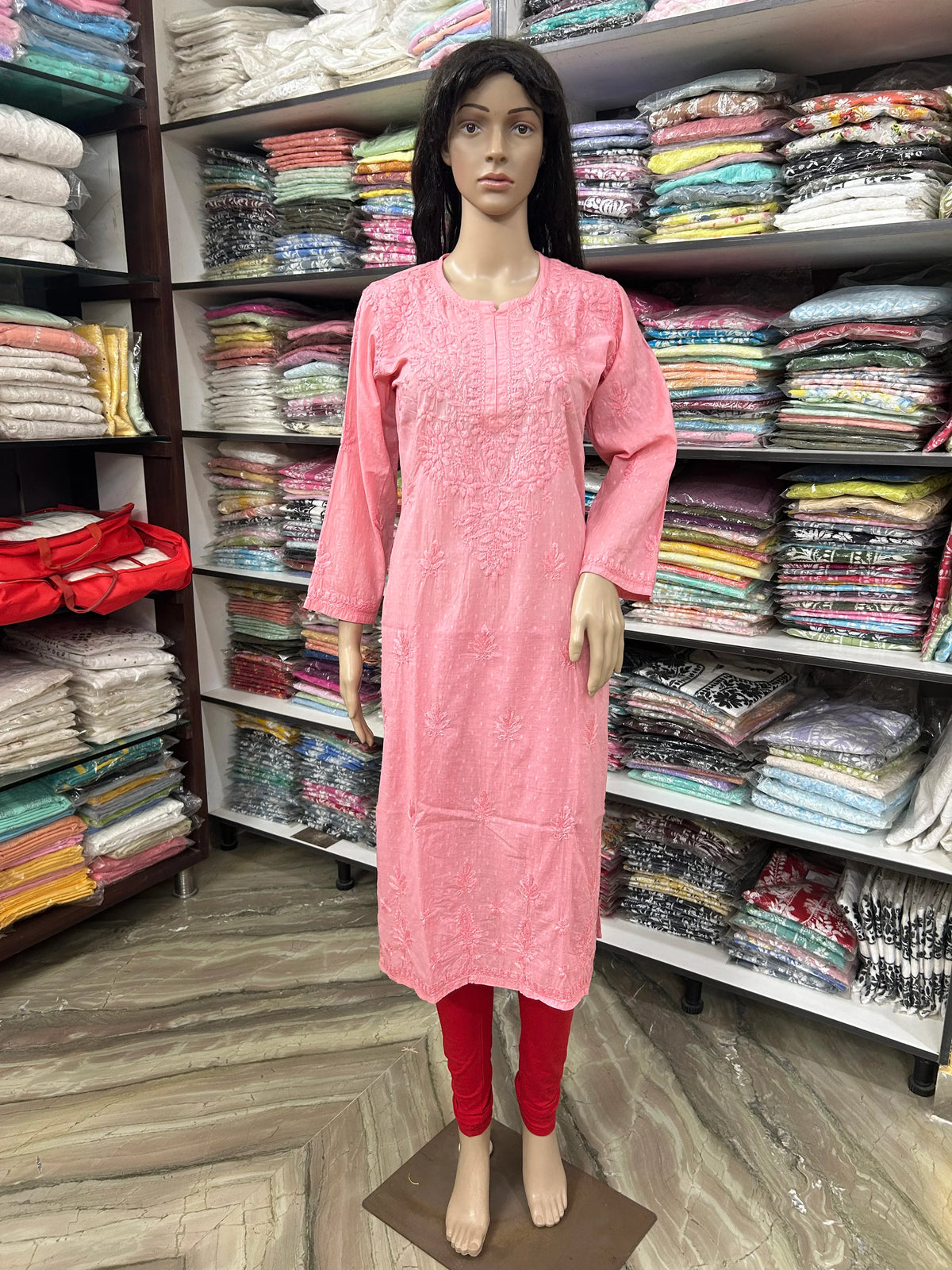A pink embroidered long kurta displayed on a female mannequin, paired with red leggings, set against a background of shelves filled with folded clothing.