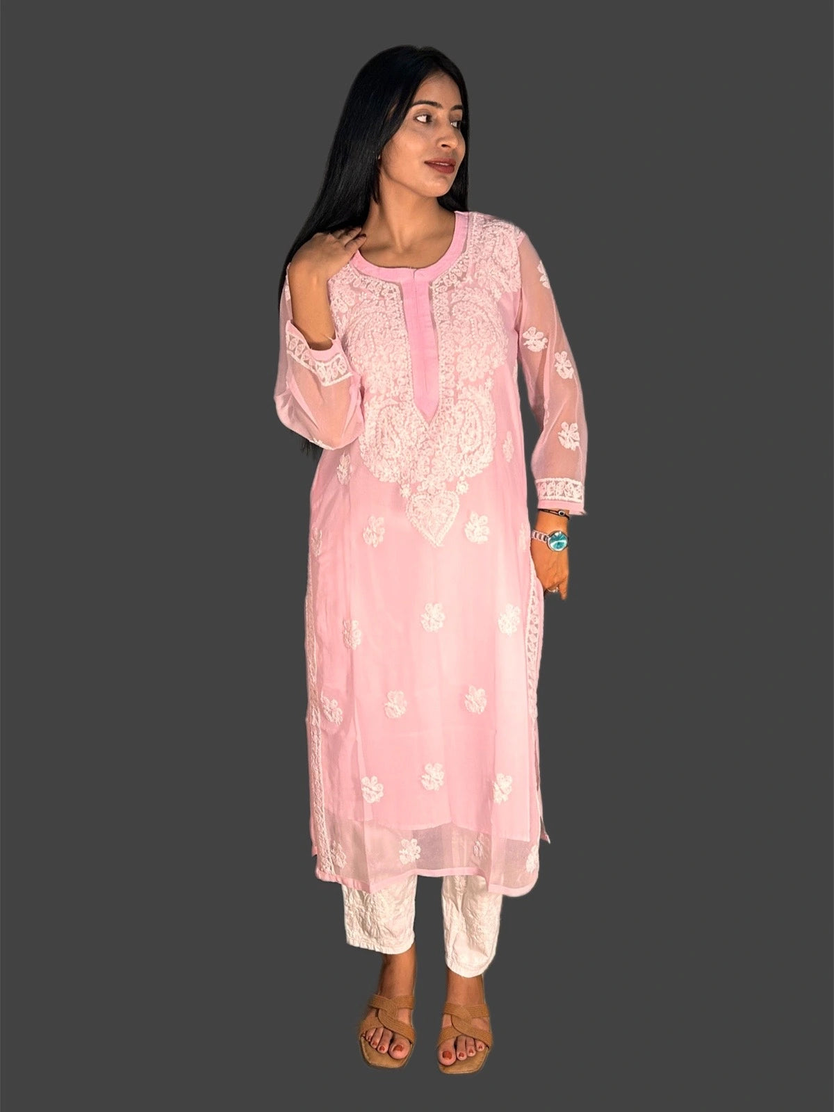 A woman modeling a light pink embroidered kurta with white floral patterns and matching white pants, standing against a solid dark gray background.