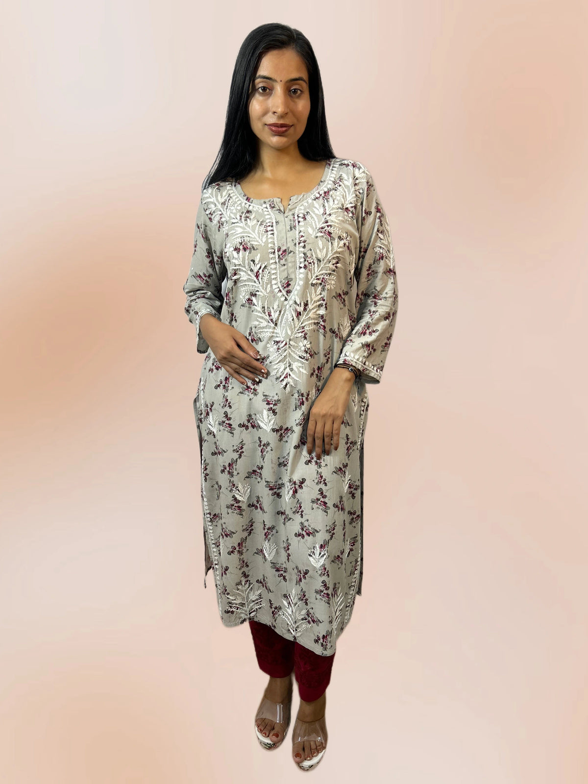 Lucknowi chikankari A woman modeling a light gray kurta with a white and dark red floral pattern, paired with dark red pants and clear high-heeled sandals, set against a soft peach gradient background.