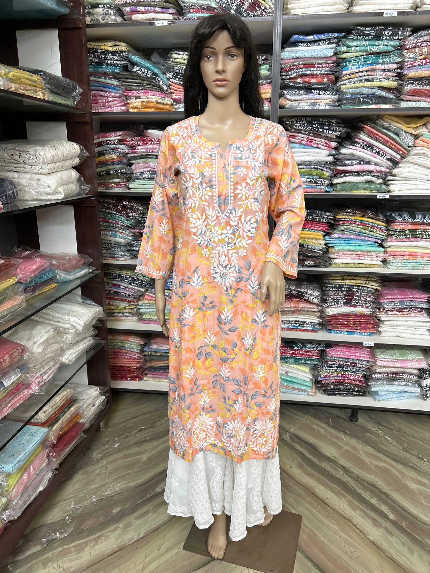 Mannequin wearing a lucknowi chikankari floral dress in a fabric store with shelves of fabric in the background.