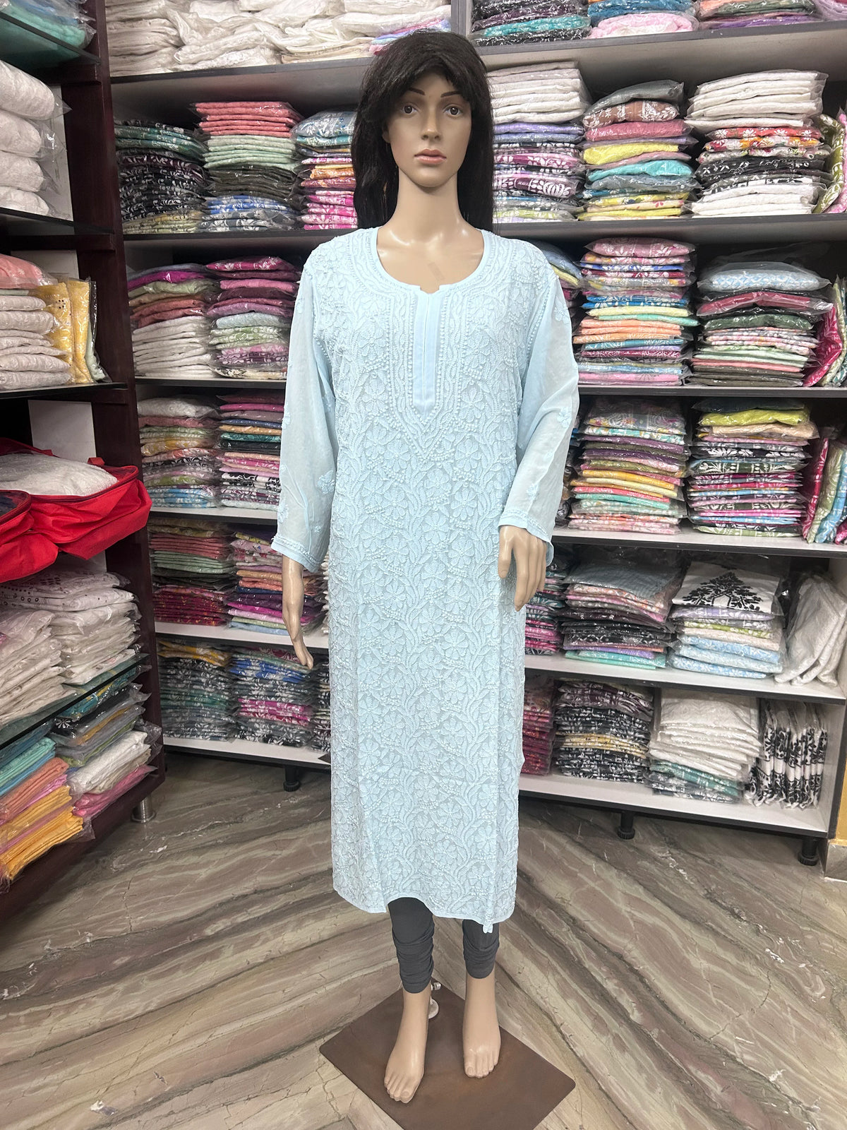A light blue long-sleeve embroidered kurta displayed on a female mannequin, standing in front of shelves filled with folded clothing.