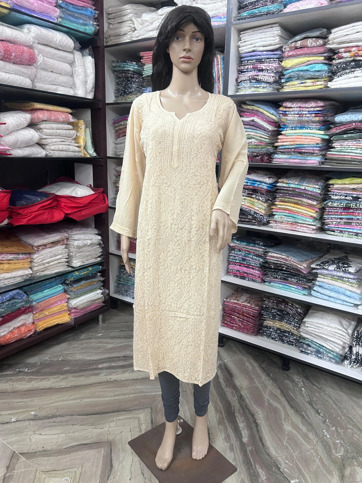 A beige long-sleeve kurta with a textured floral pattern, displayed on a female mannequin in a clothing store. The mannequin is also wearing dark gray leggings, and the background features shelves packed with folded colorful garments.