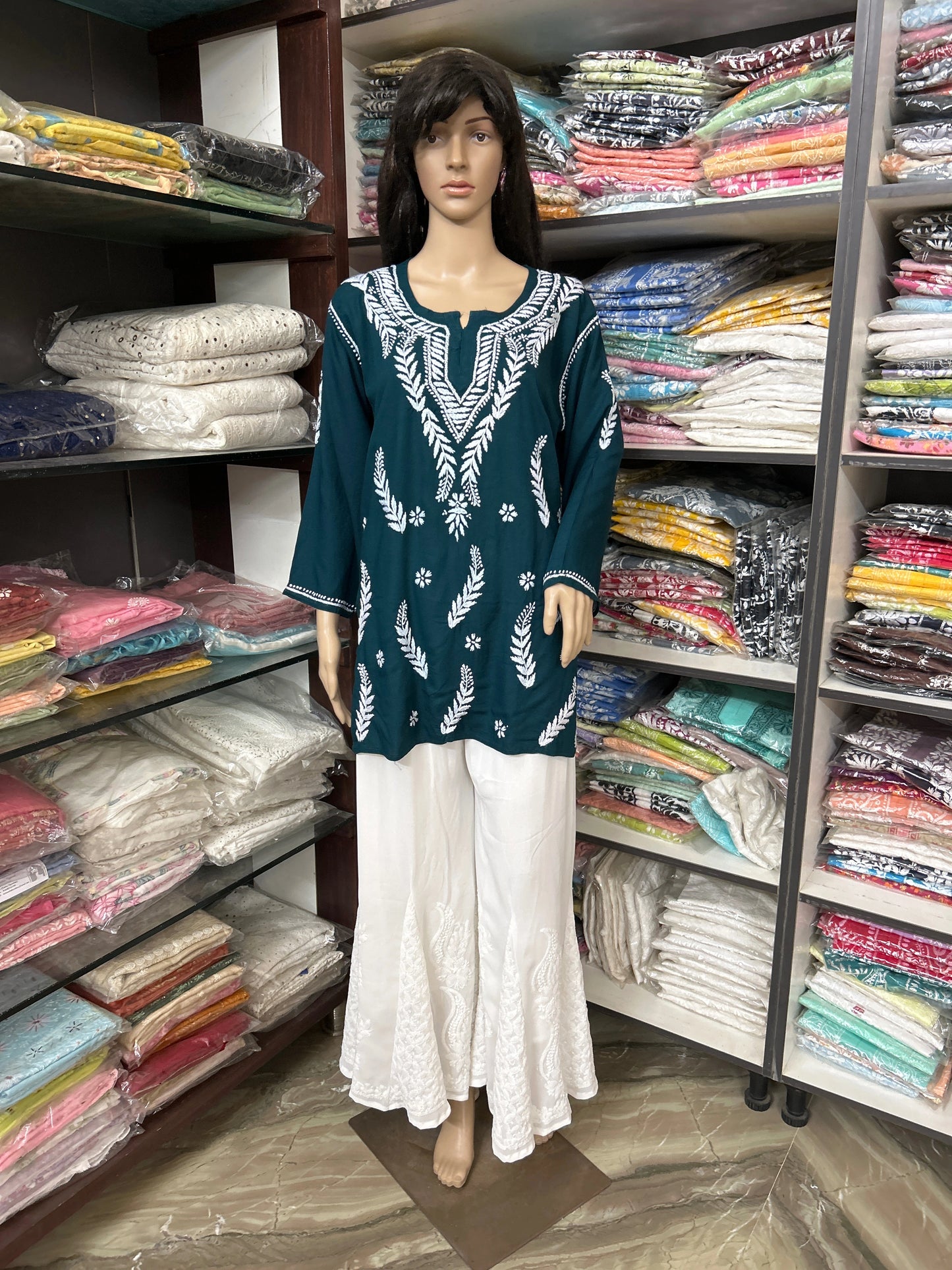 Mannequin wearing a green embroidered top and white pants in front of shelves filled with folded fabric.
