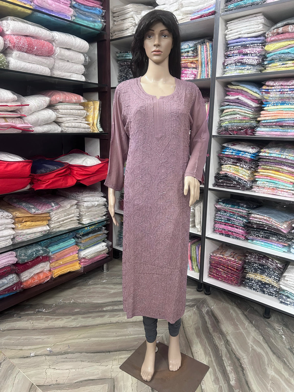 lucknowi chikankari A mannequin wearing a dusty rose pink long-sleeve kurta dress, standing in a clothing store with shelves of folded garments in the background.