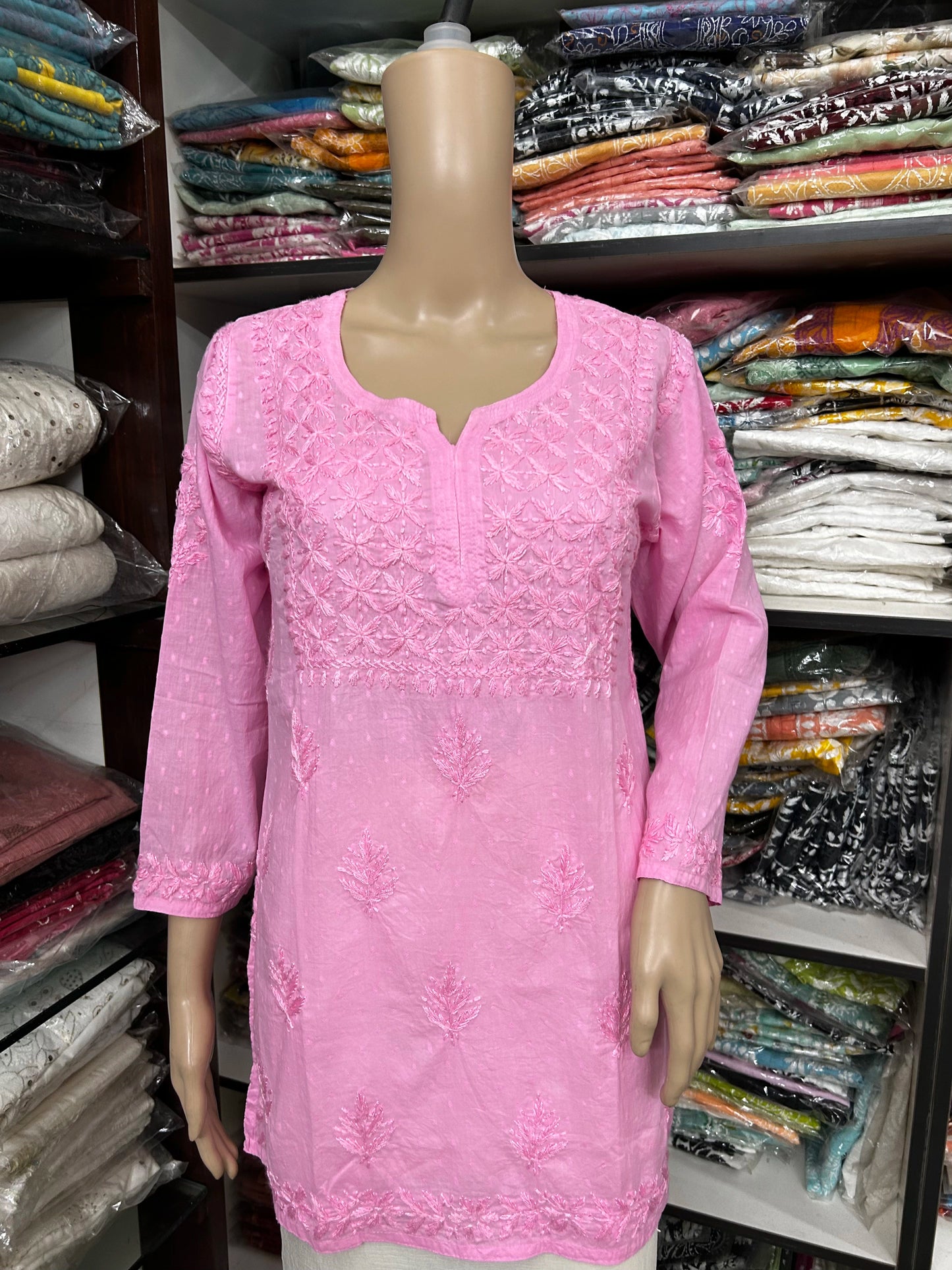 Pink embroidered kurta on a mannequin against a backdrop of fabric shelves.
