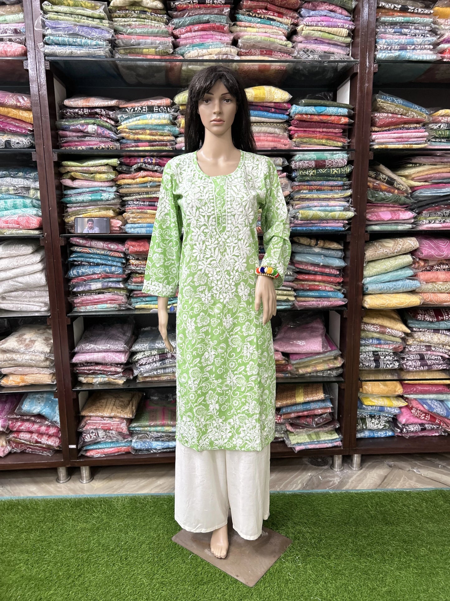 Mannequin wearing a green and white outfit in front of shelves filled with colorful fabrics.