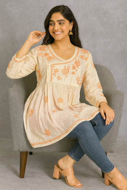 Woman sitting on a chair wearing a beige embroidered top and blue jeans.