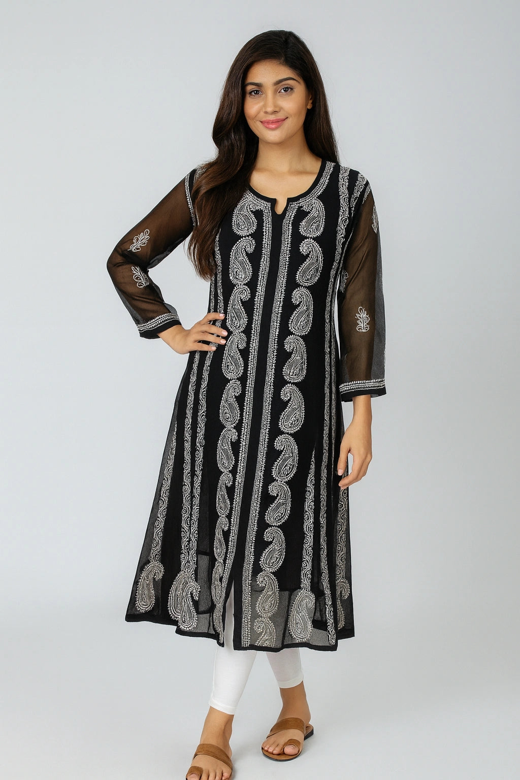 A woman modeling a black long kurta with vertical silver-gray paisley and floral embroidery, sheer black three-quarter sleeves, and white pants, standing against a light gray studio background.