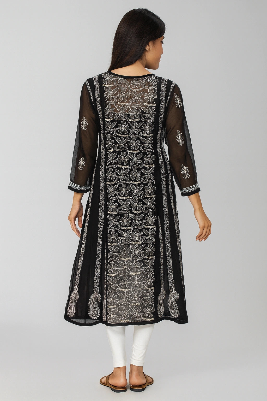 A woman viewed from the back wearing a black sheer embroidered kurta with intricate silver and white detailing, paired with white pants and brown sandals, against a light gray studio background.