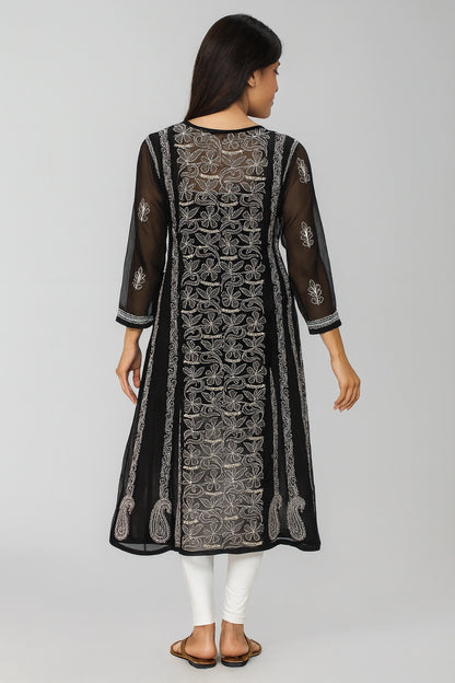 A woman viewed from the back wearing a black sheer embroidered kurta with intricate silver and white detailing, paired with white pants and brown sandals, against a light gray studio background.