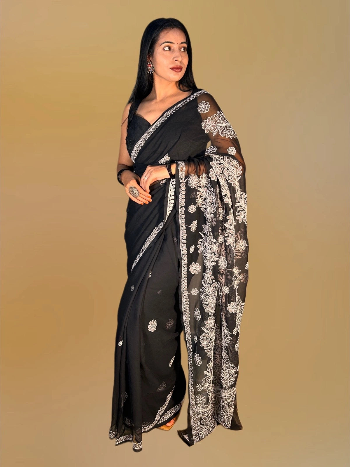 A woman modeling a black saree with intricate silver floral embroidery and border detailing, posed against a solid beige background.