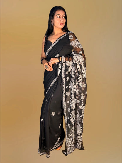 A woman modeling a black saree with intricate silver floral embroidery and border detailing, posed against a solid beige background.