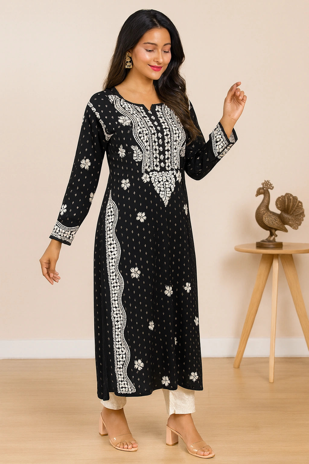 A woman modeling a black long-sleeve kurta with white floral and geometric patterns, paired with white pants and clear high-heeled sandals, in a warm beige indoor setting.