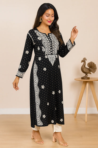 A woman modeling a black long-sleeve kurta with white floral and geometric patterns, paired with white pants and clear high-heeled sandals, in a warm beige indoor setting.