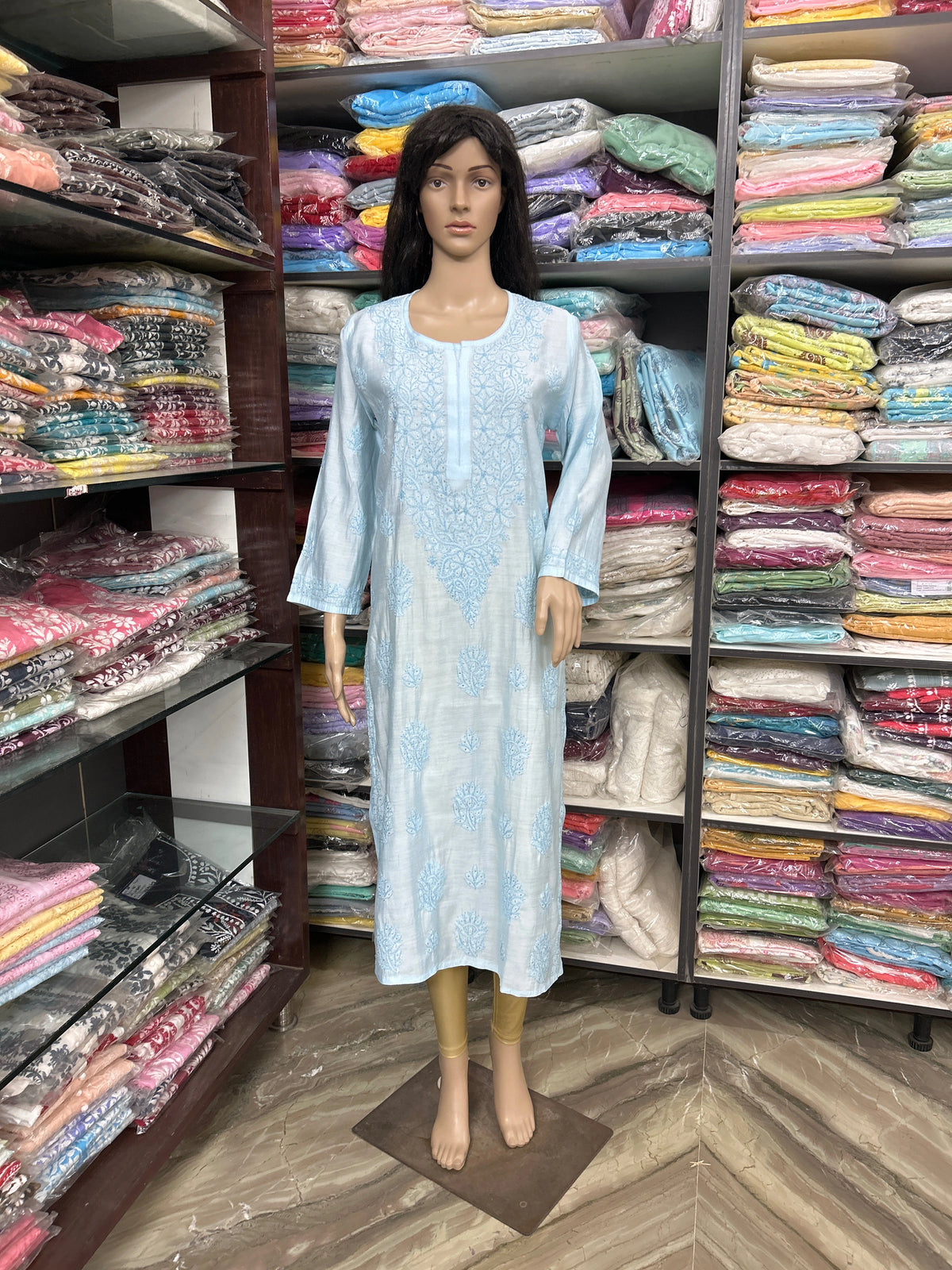 lucknowi chikankari Mannequin wearing a light blue dress in a fabric store with shelves of colorful fabrics.