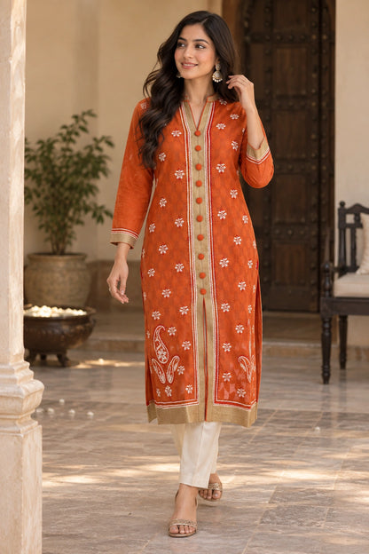 A woman models a traditional Indian outfit consisting of an orange kurta with white floral embroidery and gold buttons, paired with off-white trousers, in an elegant indoor setting with stone pillars and wooden doors.