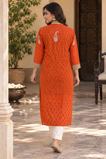 Lucknowi chikankari A woman wearing a bright orange kurta with a subtle geometric pattern, white pants, and beige shoes, shown from the back in an outdoor courtyard setting