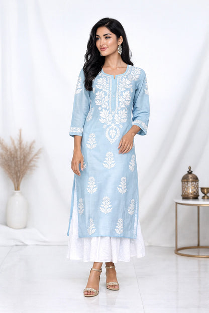 A woman models a light blue kurta with white floral embroidery, paired with a white flared underskirt and beige sandals, set against a white draped studio background.