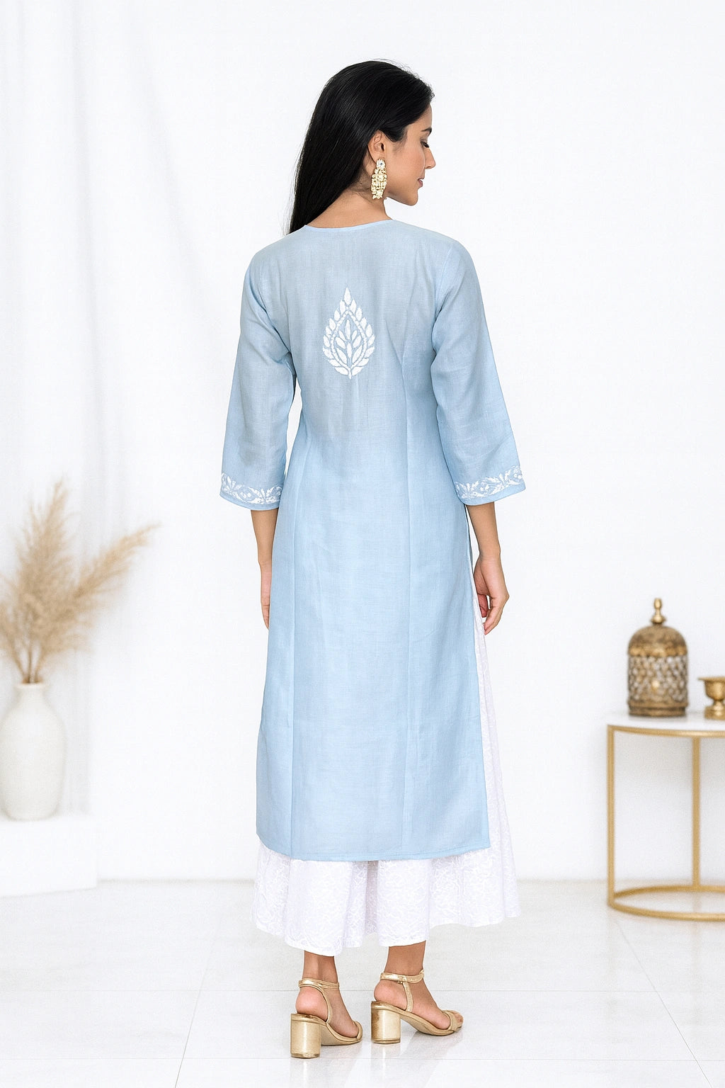 A woman stands with her back to the camera wearing a light blue kurta with white embroidery on the back and white side slits, paired with white pants and gold sandals in a bright studio setting.