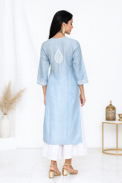 A woman stands with her back to the camera wearing a light blue kurta with white embroidery on the back and white side slits, paired with white pants and gold sandals in a bright studio setting.