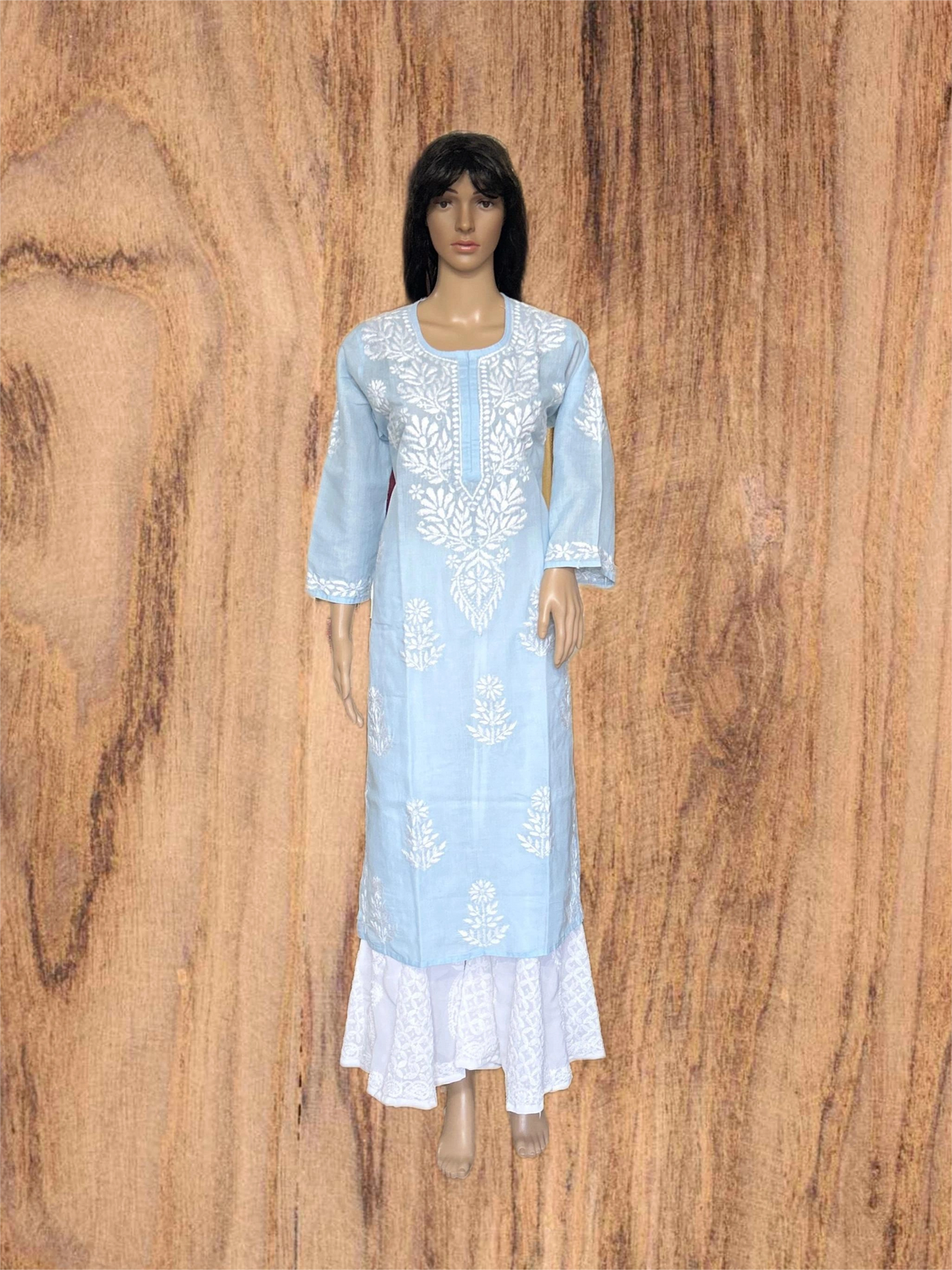 A light blue long kurta dress with white floral embroidery and a white lace hem, displayed on a female mannequin against a wood grain background.