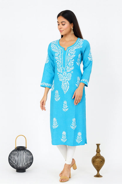A woman modeling a bright turquoise blue kurta with white floral embroidery, styled with white pants and beige heels, standing against a white studio background with decorative props.