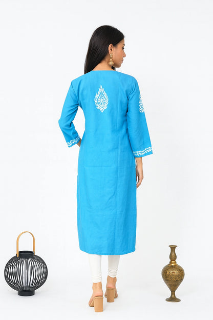 A woman is shown from the back wearing a bright blue kurta with white embroidery on the back and sleeve cuffs, paired with white pants and beige heels, set against a plain white background with decorative props.