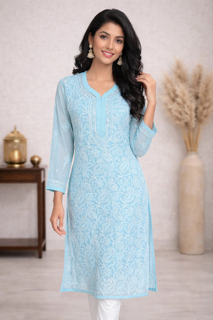 A woman models a blue embroidered kurta with three-quarter sleeves and white leggings in an indoor setting with a wooden cabinet.