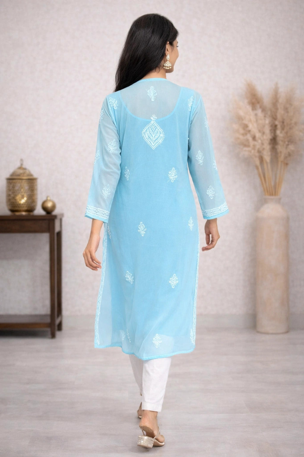 A woman viewed from behind wearing a blue long kurta with three-quarter sleeves and a subtle embroidered motif on the back, paired with white leggings in an indoor setting.