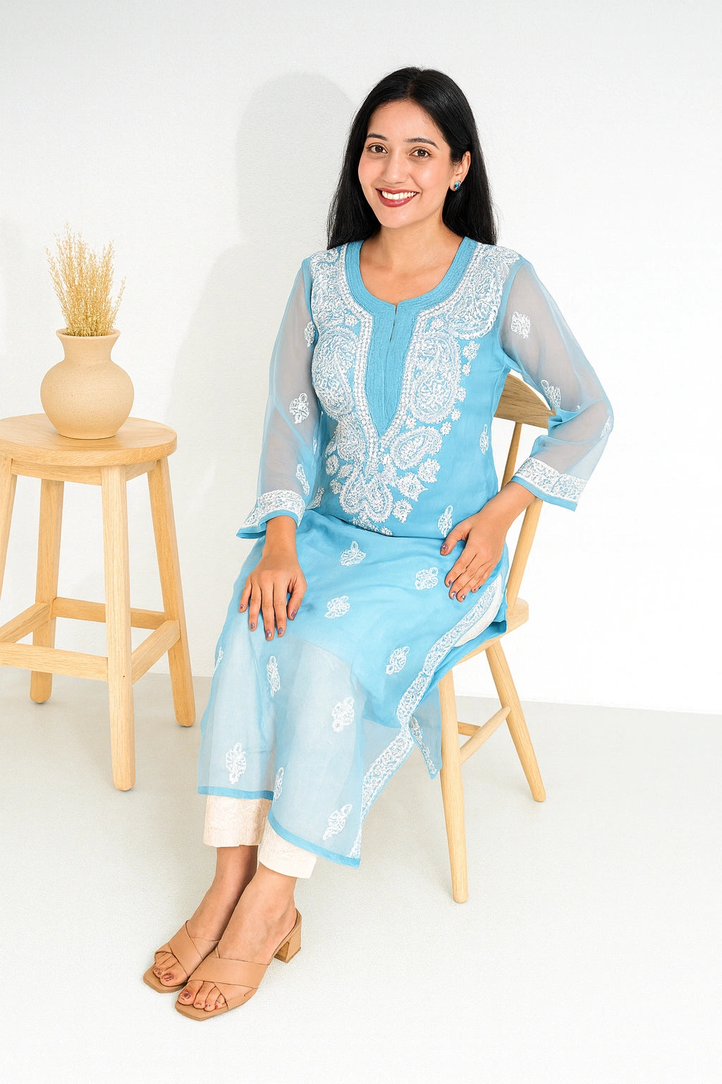 A woman wearing a light blue embroidered kurta with white floral patterns and sheer sleeves, seated on a wooden chair next to a small wooden stool with a vase of dried grass.