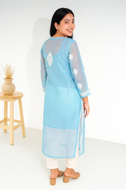 A woman wearing a light blue sheer kurta with white floral embroidery on the back and sleeves, paired with white pants and beige sandals, standing in a bright studio setting.