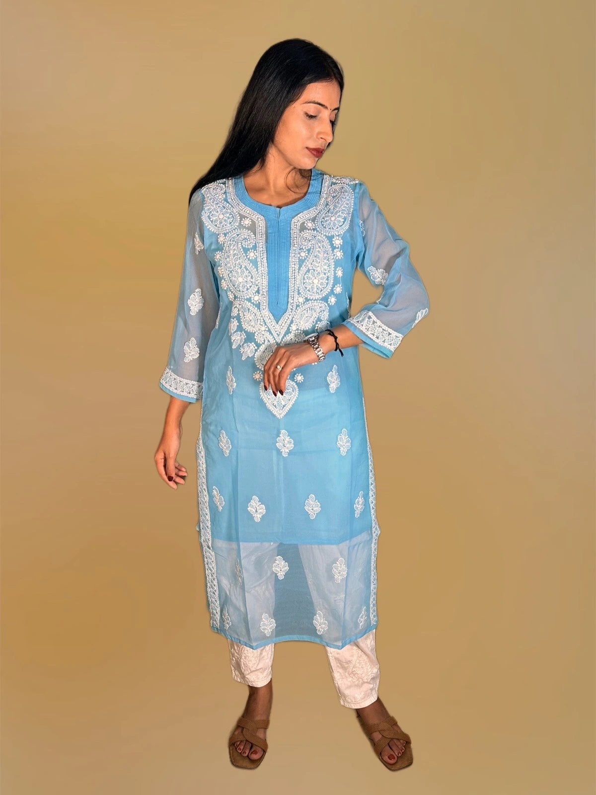 A woman modeling a light blue embroidered kurta with white lace details, paired with white pants and brown sandals, set against a solid beige background.