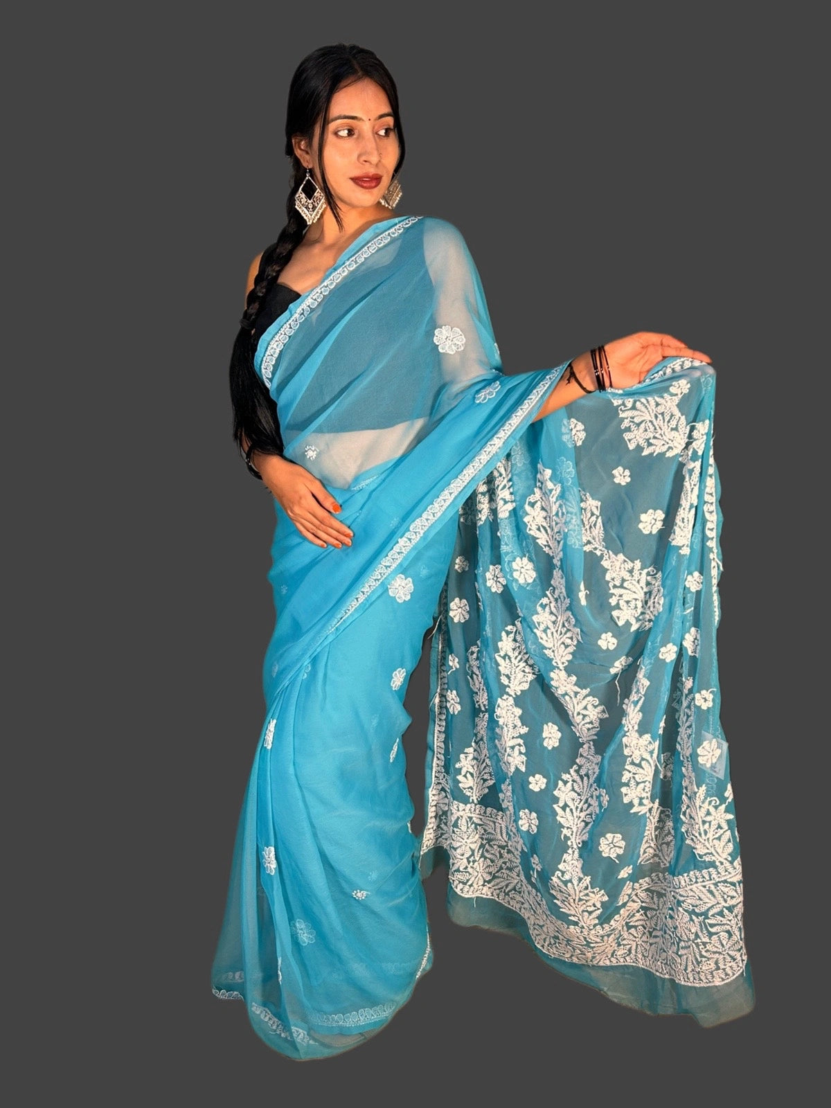 A woman modeling a light blue saree with white floral embroidery and a black blouse, posed against a solid dark gray background.