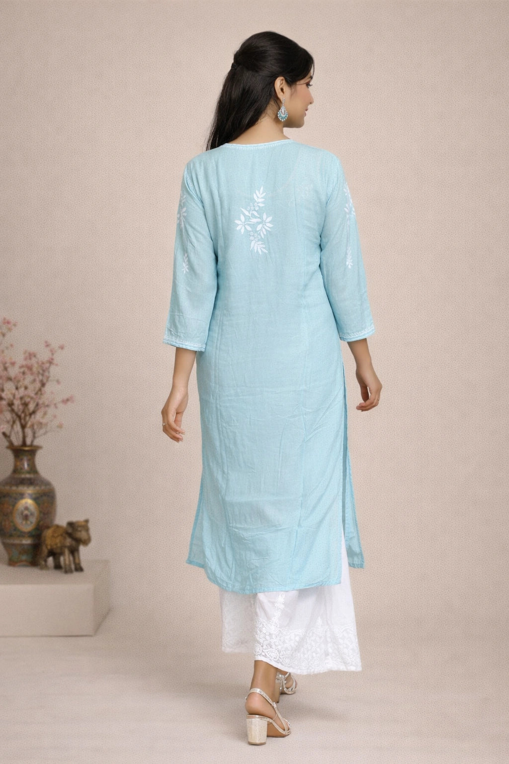A woman is shown from the back wearing a light blue cotton kurta with white floral embroidery on the upper back, paired with white pants and beige sandals, set against a neutral beige background.