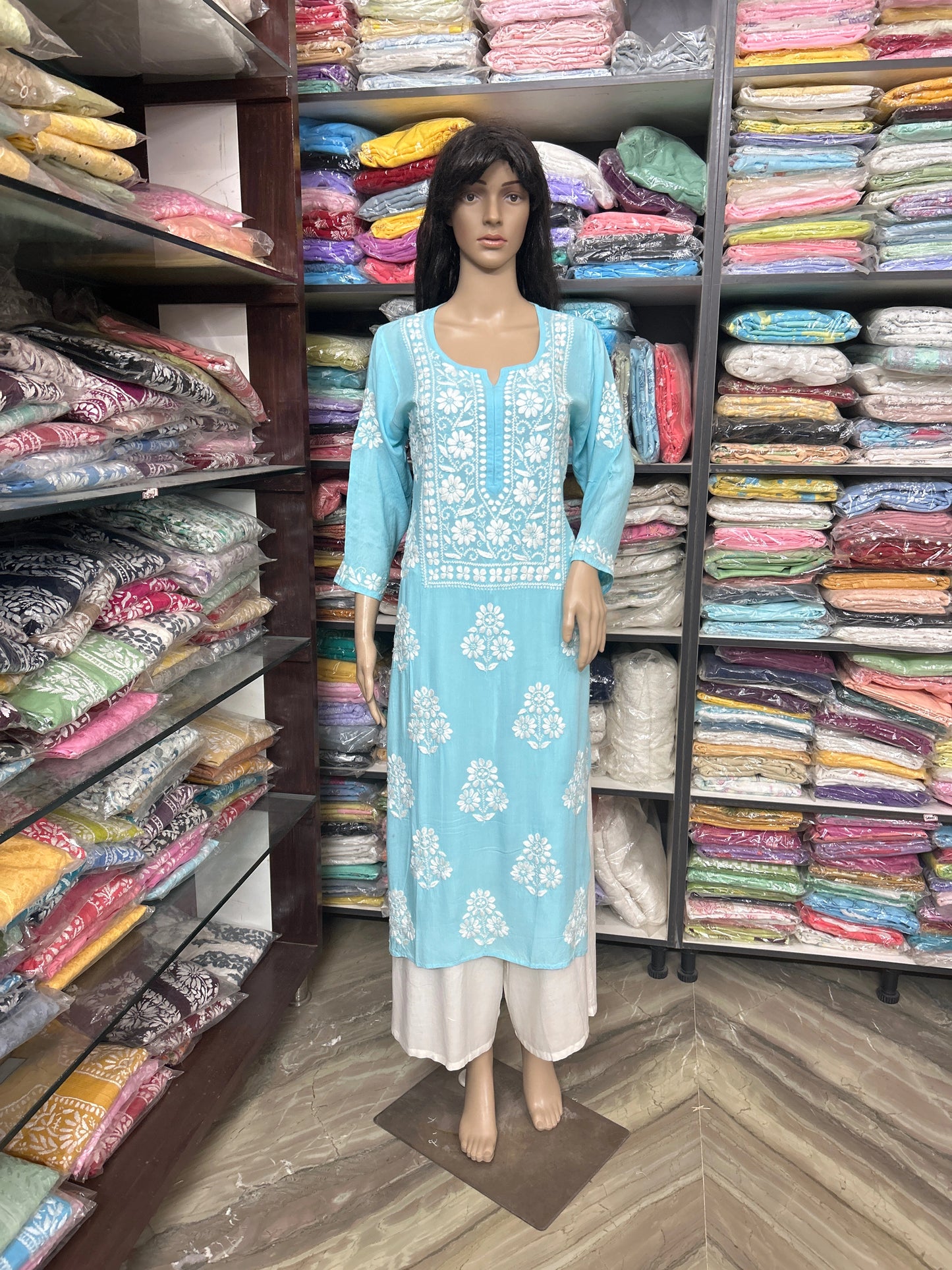 Mannequin wearing a blue embroidered dress in a fabric store with shelves of colorful fabrics.