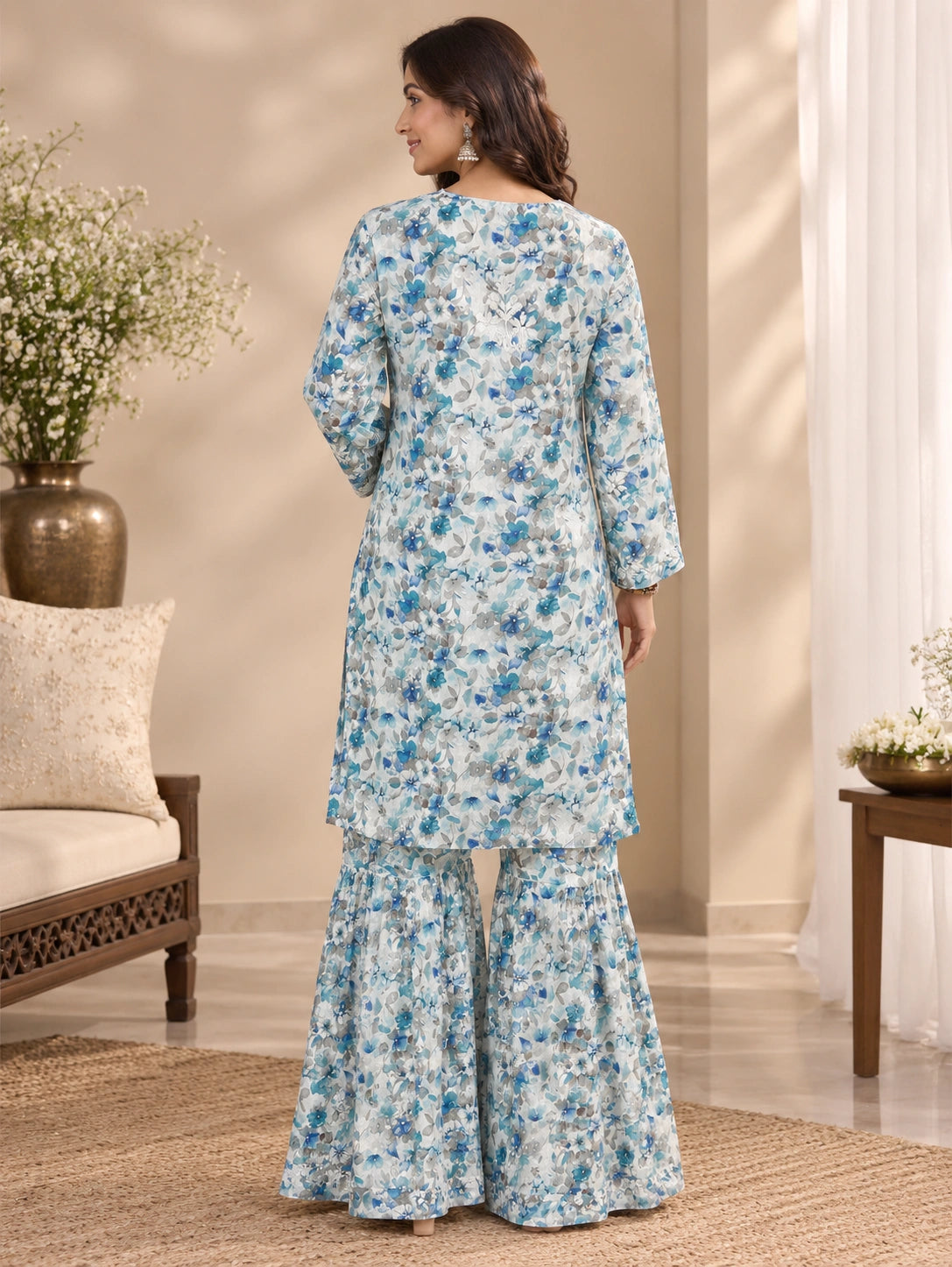 Lucknowi chikankari A woman stands with her back to the camera wearing a light blue and white floral printed kurta set with matching flared pants in a warm, decorated indoor setting.