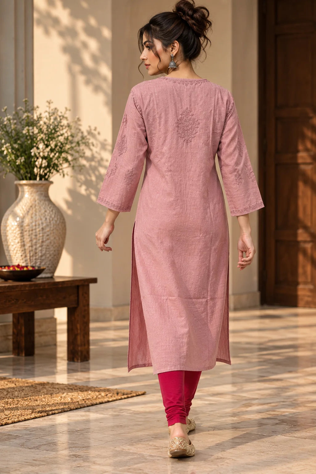 A woman wearing a dusty pink kurta with three-quarter sleeves and a subtle embroidered motif on the upper back, paired with magenta leggings and beige flat shoes, walking in a sunlit indoor setting.