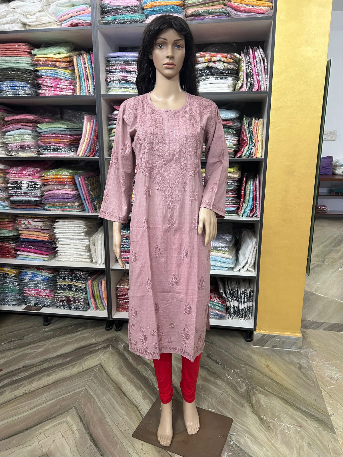 A pink embroidered kurta displayed on a female mannequin, paired with red leggings, set against a background of shelves filled with folded colorful fabrics.
