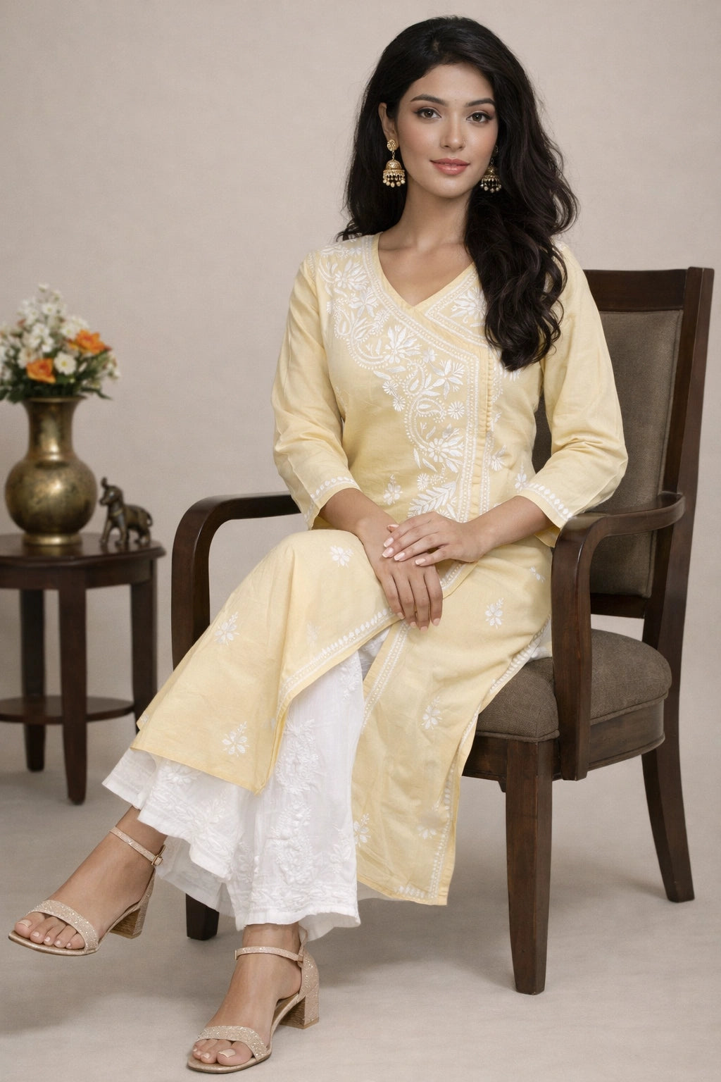 A woman seated in a wooden armchair wearing a light yellow embroidered kurta with white flared pants, styled in beige heeled sandals against a neutral beige studio background.