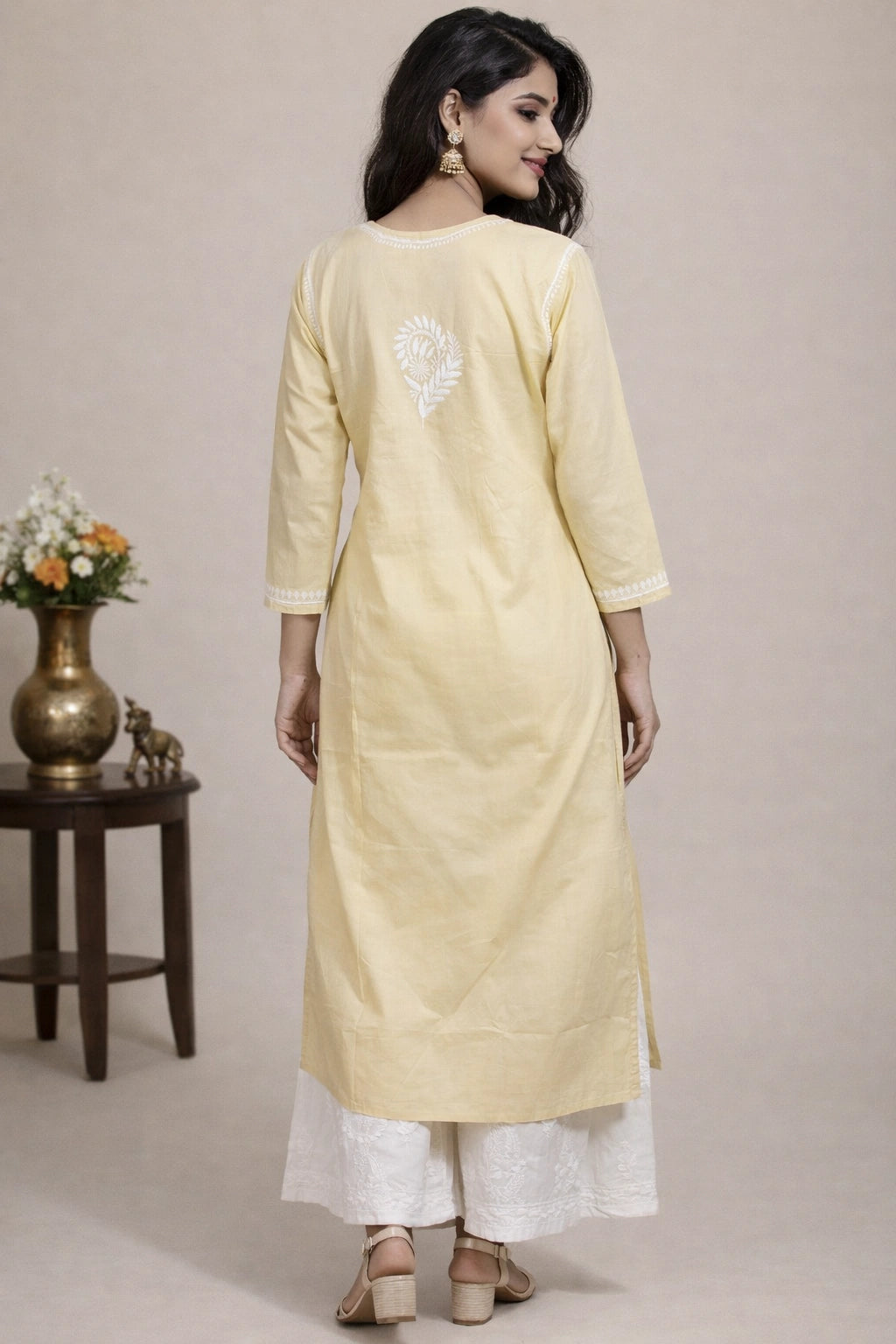 A woman wearing a pale yellow three-quarter sleeve kurta with white embroidery on the back, paired with white palazzo pants, shown from a rear three-quarter view in a studio setting.