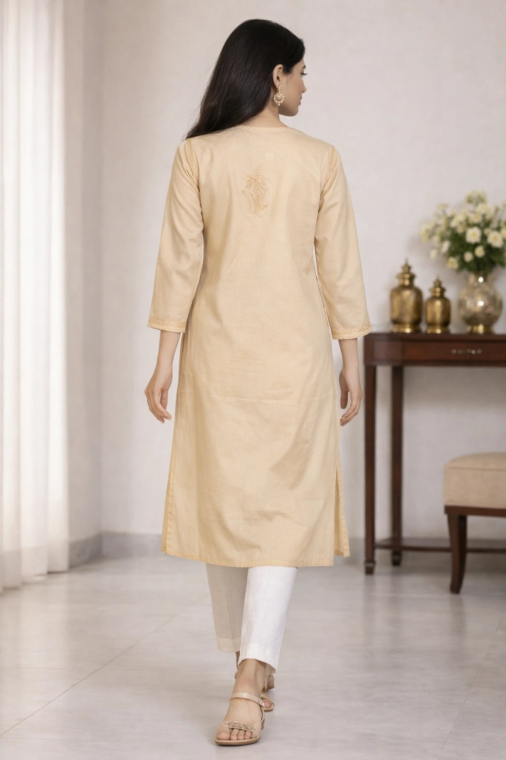 Lucknowi chikankari A woman is shown from behind wearing a light beige kurta with white pants and beige sandals in an indoor setting.