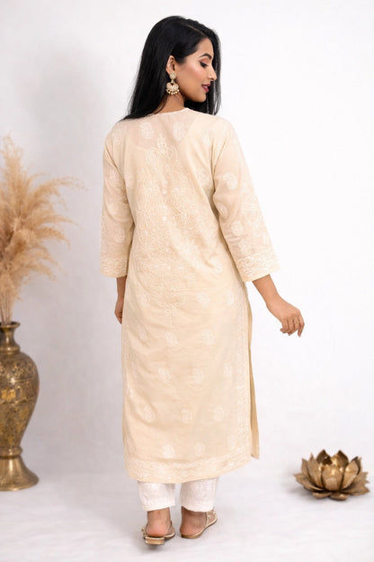 A woman wearing a light beige embroidered kurta with white pants, shown from the back in a studio setting with decorative dried grass in a vase and a gold lotus ornament.