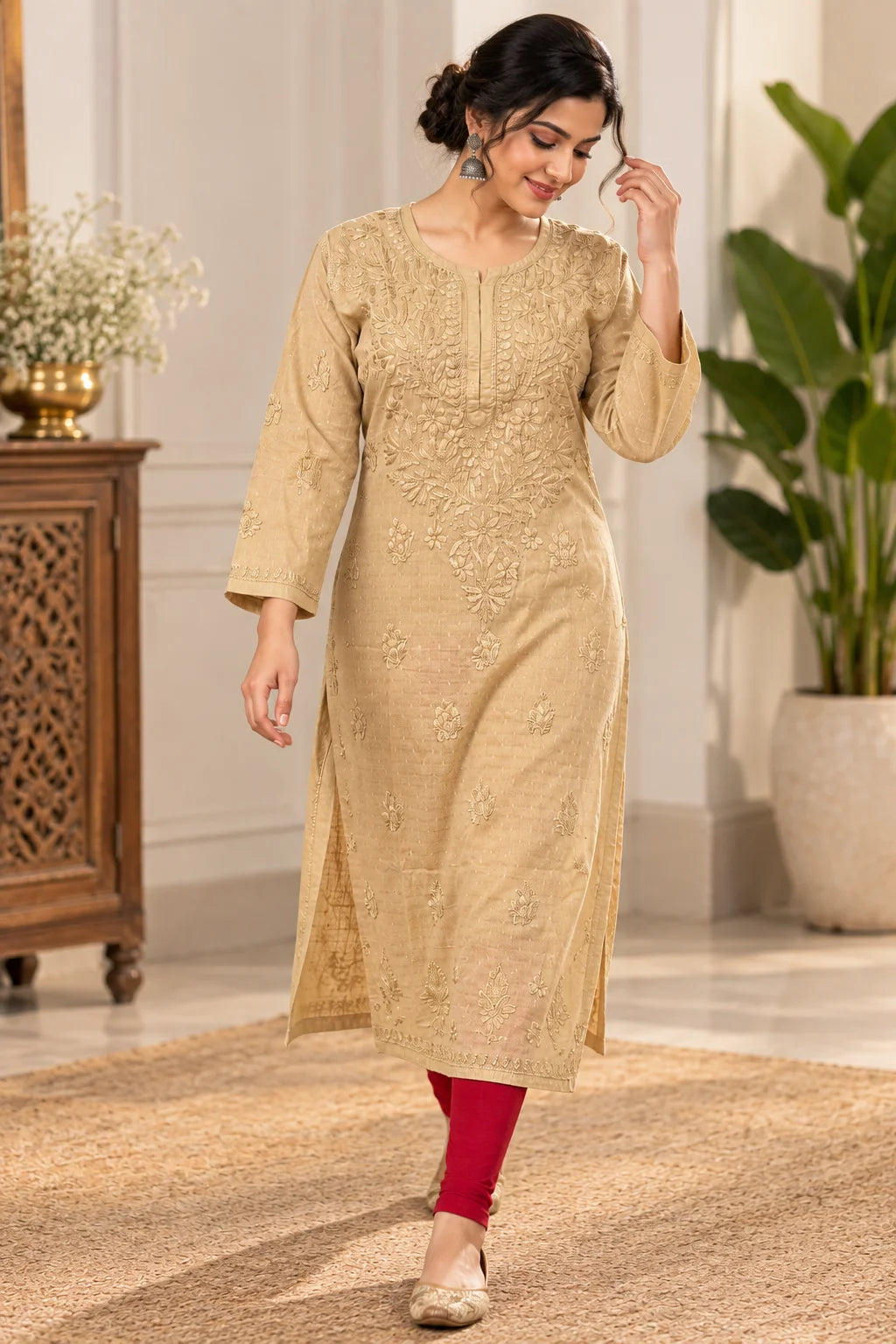 A woman models a beige embroidered kurta with three-quarter sleeves and red leggings in an indoor setting with a wooden cabinet and potted plants.