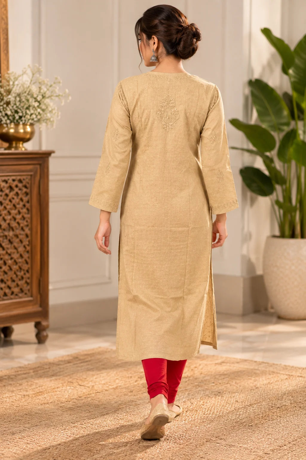 A woman viewed from behind wearing a beige long kurta with three-quarter sleeves and a subtle embroidered motif on the upper back, paired with bright red leggings and beige flat shoes in an indoor setting.