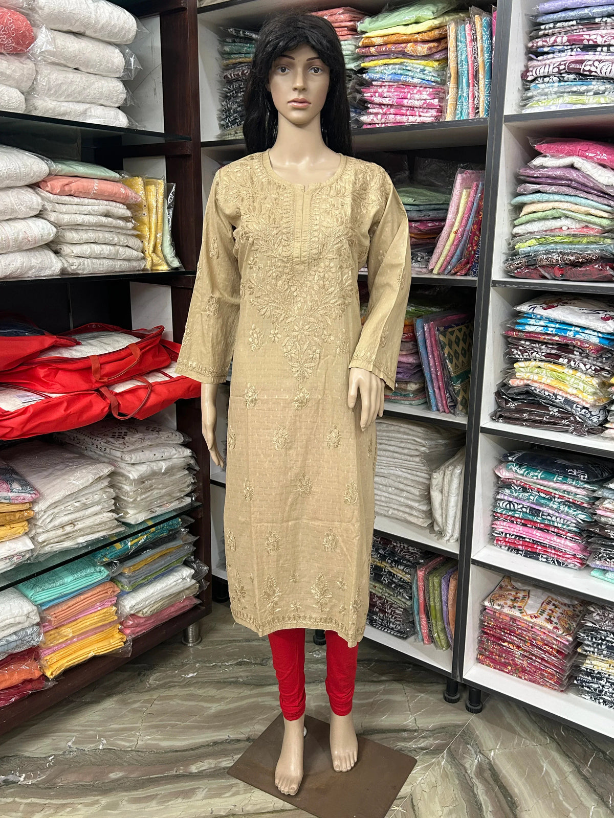 A beige embroidered kurta displayed on a female mannequin, paired with red leggings, set inside a clothing store with shelves of folded garments in the background.